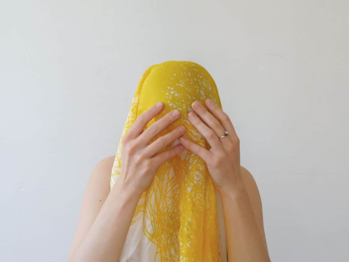 video still of person with head covered by a yellow and white scarf, hands to their face.