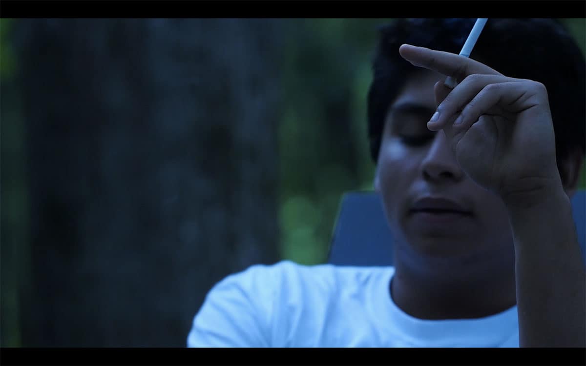 a video still of a young man wearing a white shirt sitting outside in dusky light holding a cigarette