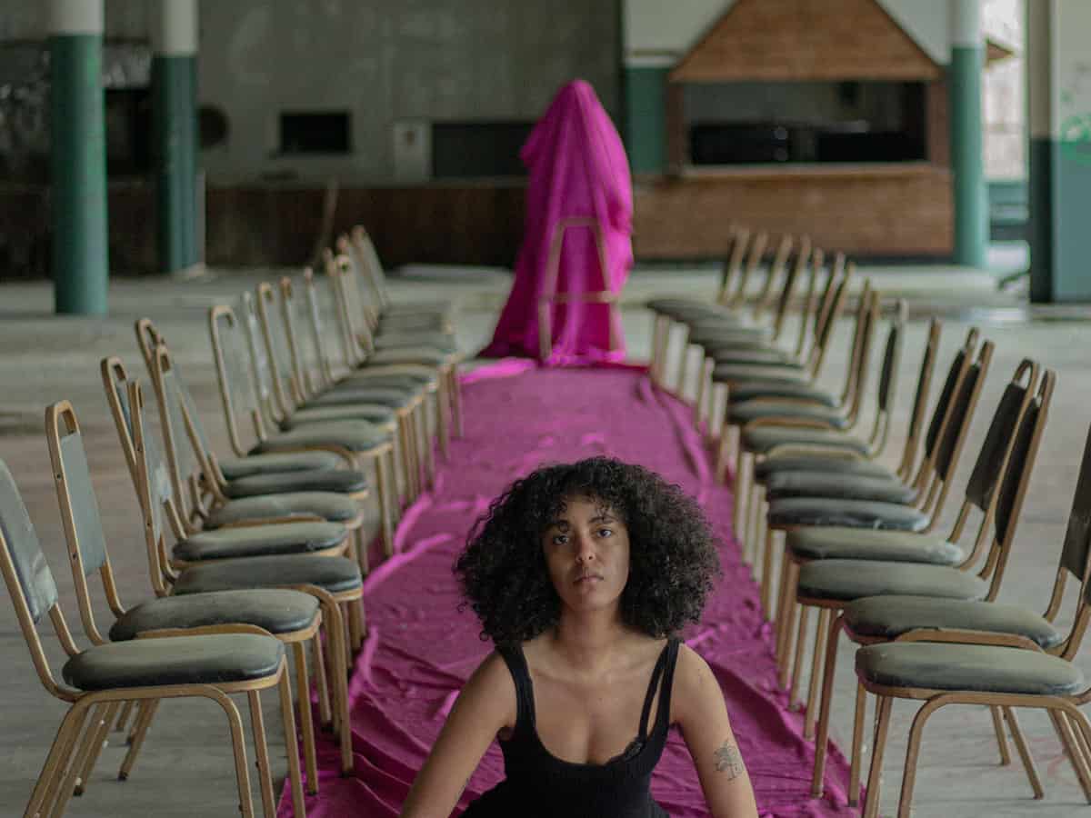 video still of woman sitting on a purple carpet with a figure behind her covered in purple fabric