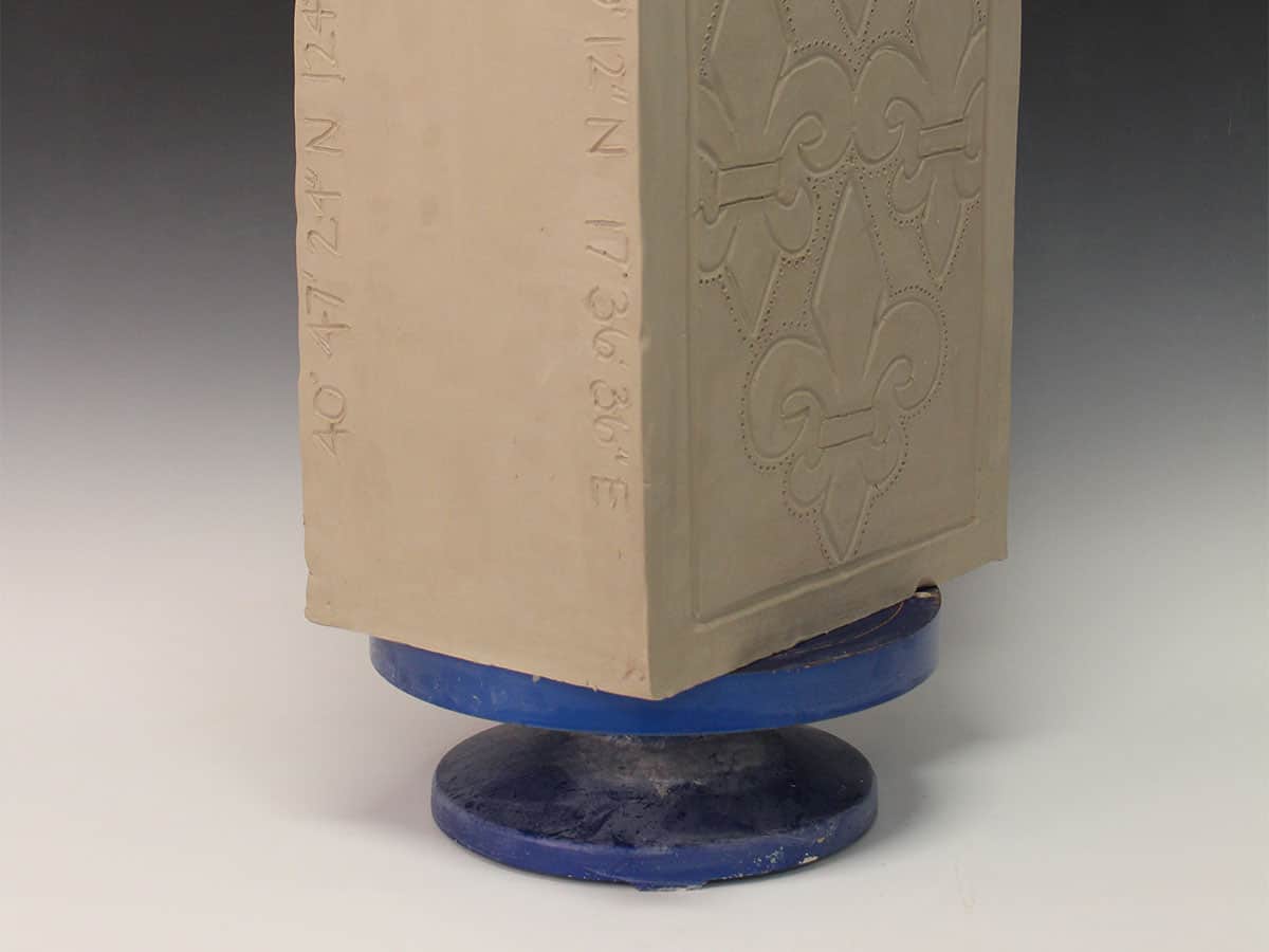box-shaped ceramic object on a blue pedestal. one side is inscribed with latitudes and longitudes and the other is inscribed with a fleur de lis pattern.