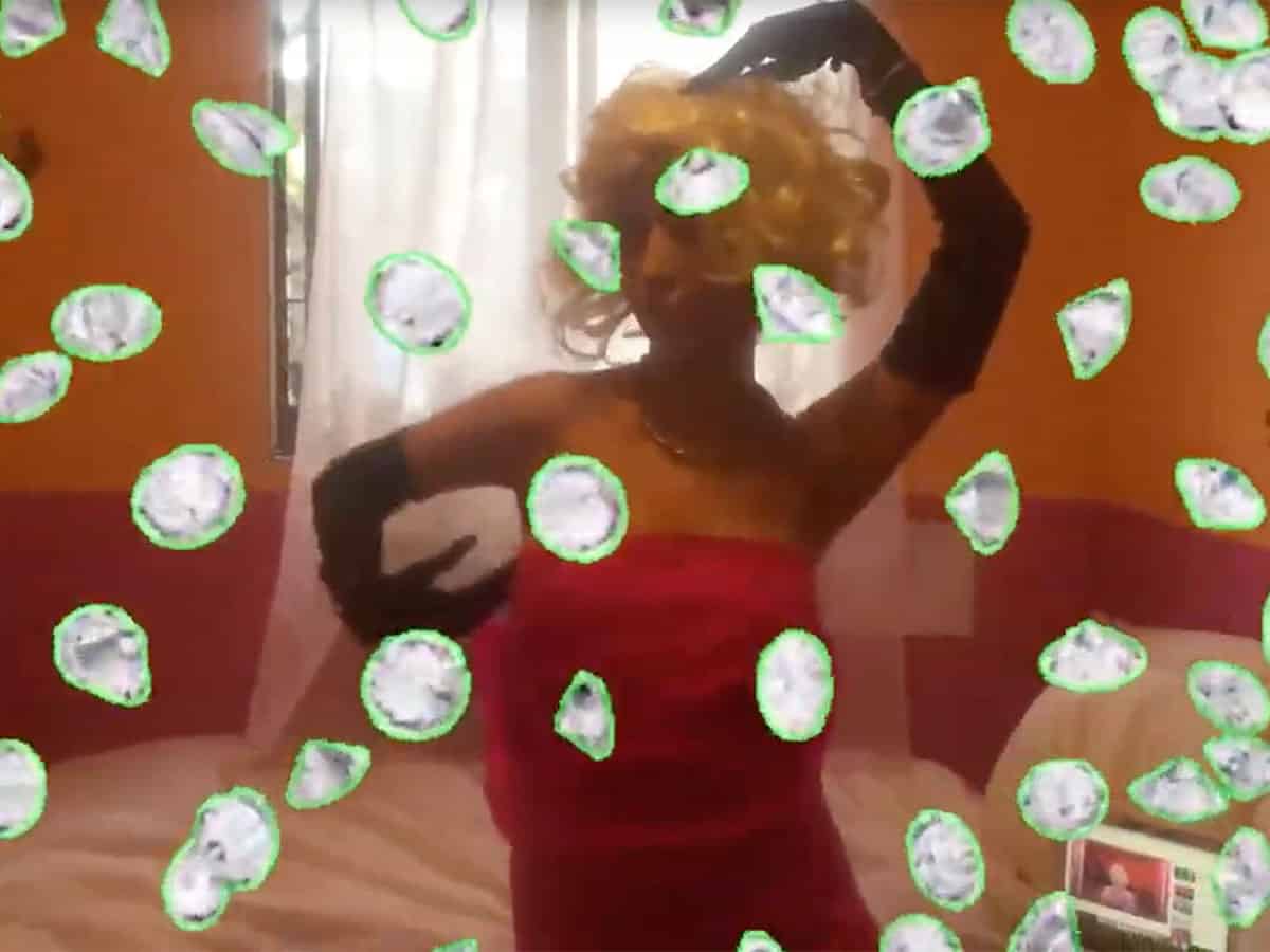 video still of woman in red dress, wearing long black gloves and a blond wig. overlaid with green-outlined falling diamonds.